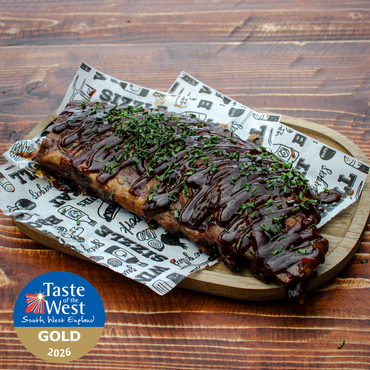 A platter of BBQ ribs topped with barbecue sauce and chopped green herbs. A Taste of the West South West England Gold 2026 badge is in the lower-left corner.