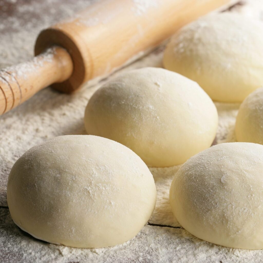 Four smooth, flour-dusted dough balls arranged on a floured surface with a wooden rolling pin in the background.