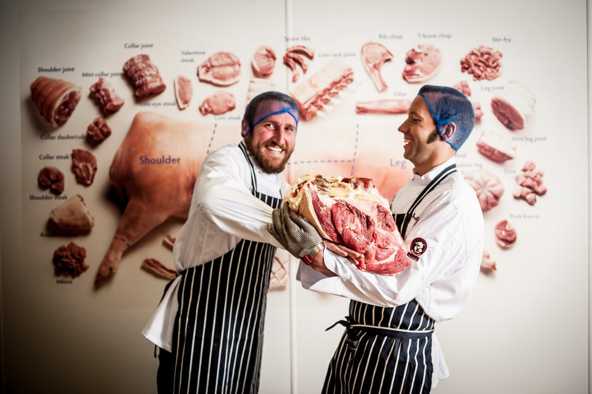 Two men in white uniforms and black-and-white striped aprons hold a large raw beef joint in front of a wall covered with labelled meat anatomy diagrams.