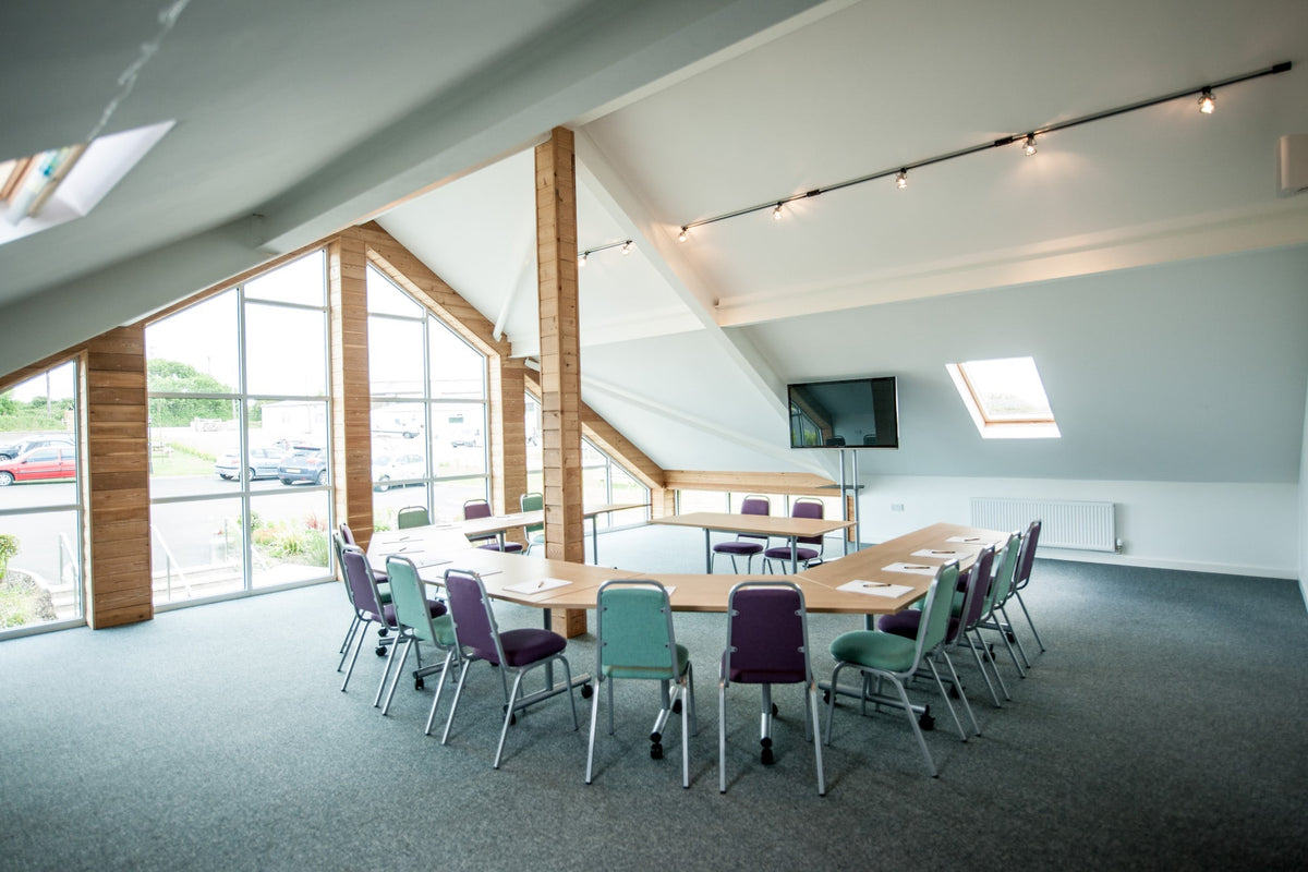 A modern meeting room with a large U-shaped wooden conference table and large monitor surrounded by teal, purple, and green chairs, set in a spacious room with a sloped ceiling, wood columns, and large windows.