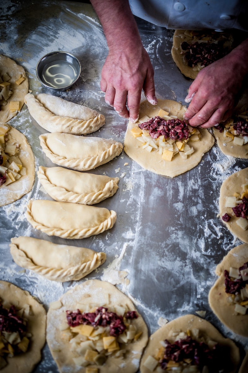 Hands preparing Cornish pasties on a floured metal baking surface, with several filled and sealed pasties arranged around them.