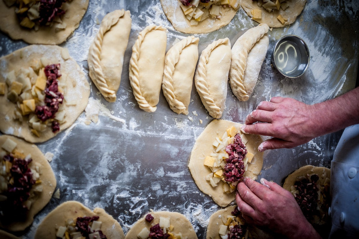 Hands preparing Cornish pasties on a floured metal baking surface, with several filled and sealed pasties arranged around them.