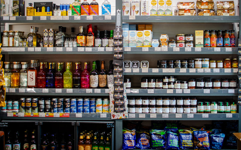 A grocery store shelf stocked with an assortment of packaged foods and beverages, including bottled spirits, canned drinks, jars, and bags of chips.