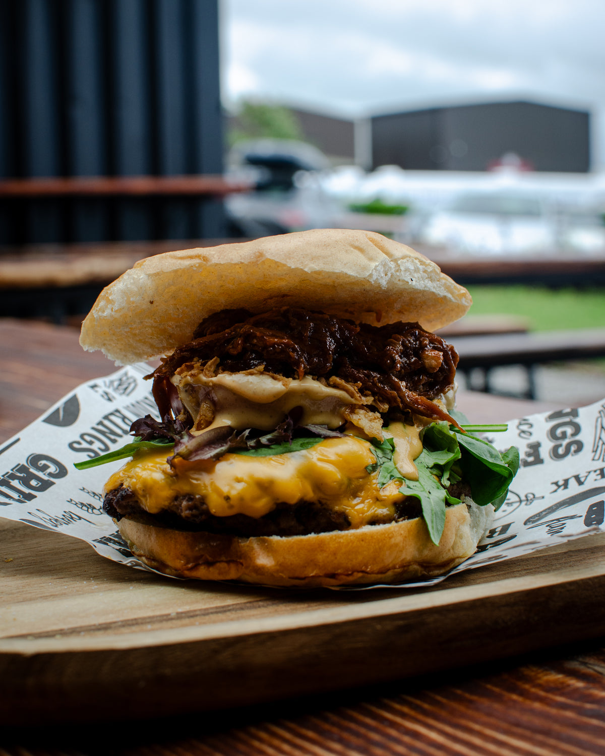 A large gourmet burger with a beef patty, BBQ pulled pork, melted cheese, crispy fried onions, burger sauce, fresh greens, and a toasted bun.