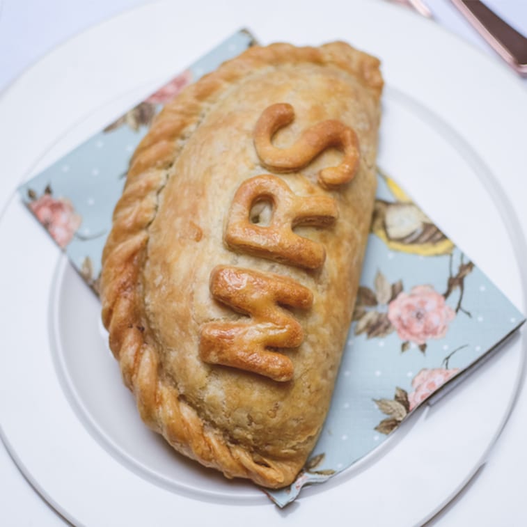 A baked Cornish pasty with 'MRS' in decorative letters on top, resting on a floral napkin on a white plate.
