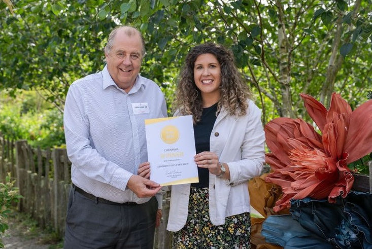 A man and a woman standing outdoors in front of green trees, holding a certificate. 