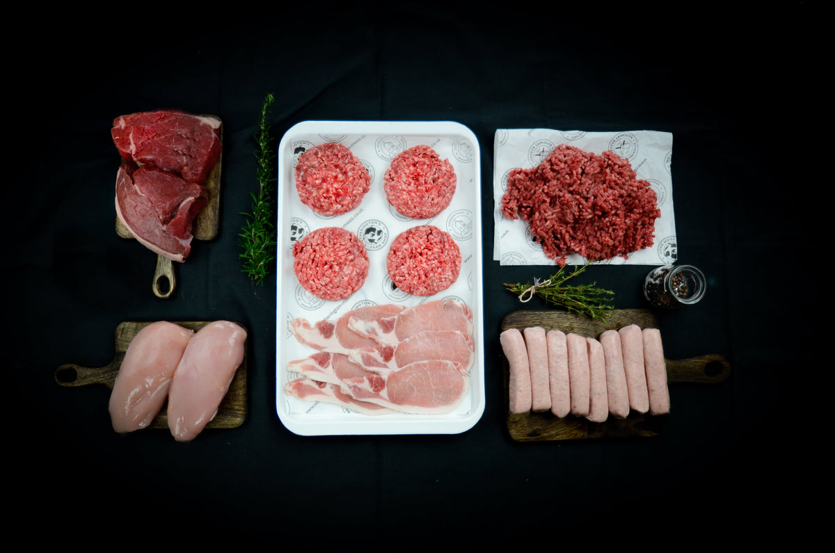 Raw meat ingredients arranged on a black background: two raw chicken breasts, four bacon rashers, a tray with four raw burger patties and a tray of ground meat, and a board of sausages, with small sprigs of rosemary and cuts of steak.