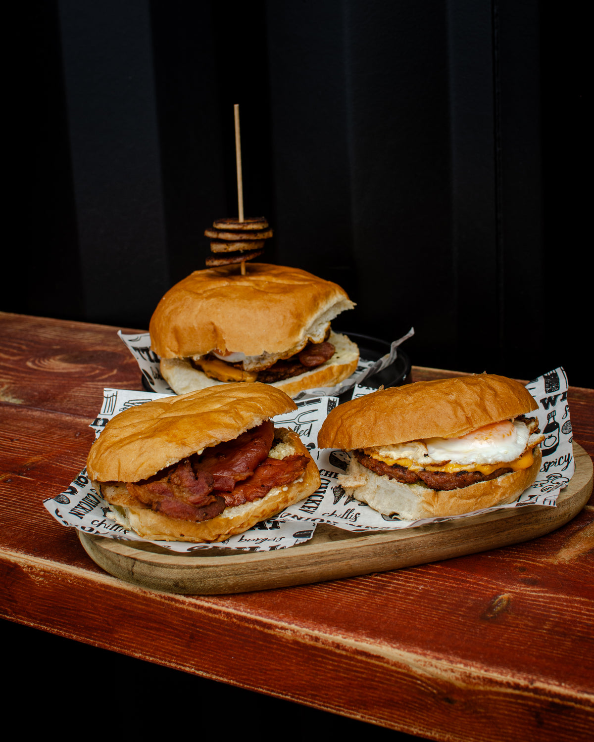 Three gourmet breakfast sliders on a wooden board, featuring toasted buns, sausage, bacon, cheese, hogs pudding, hash browns and eggs.