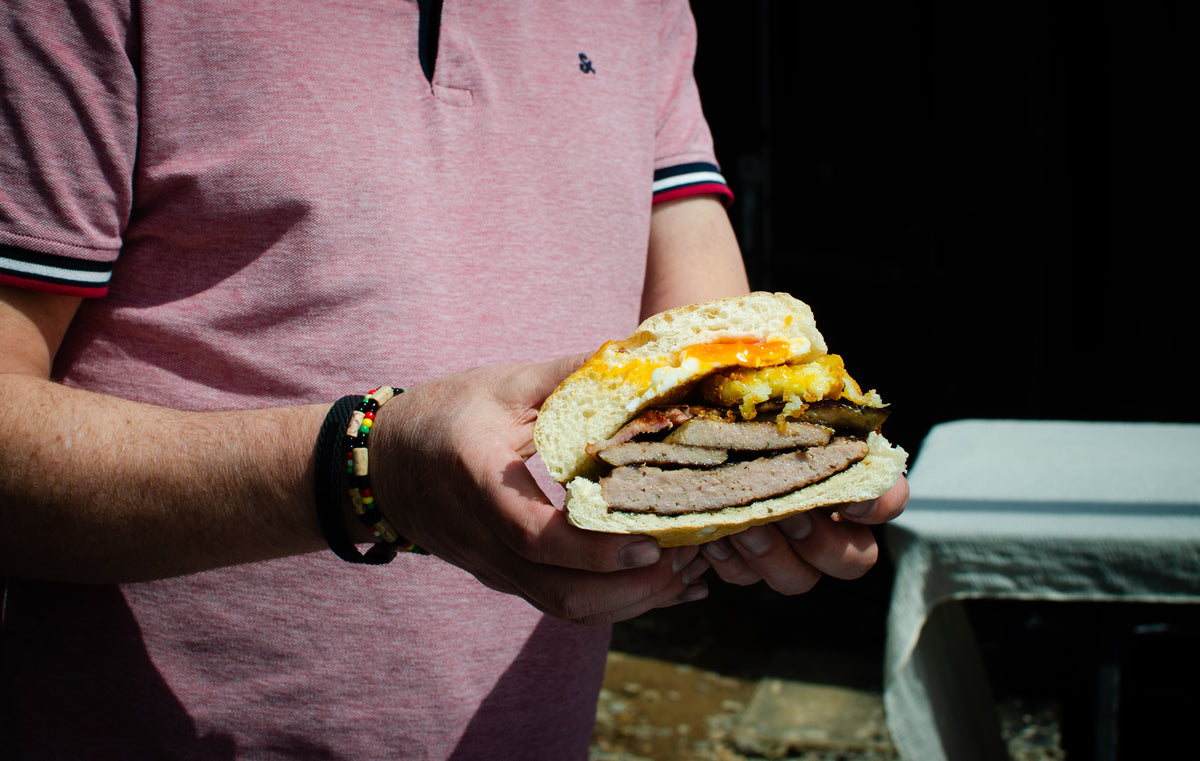 A close-up of a person holding a large breakfast bap with a sausage patty, bacon, hogs pudding, melted cheese, and egg and hash browns