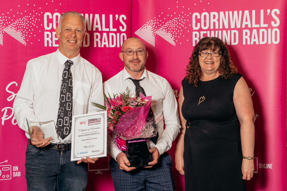 Three people standing together in front of a bright pink Cornwall's Rewind Radio backdrop, with the centre person holding bouquet. The person on the left is holding a framed award for Family Business of the Year.