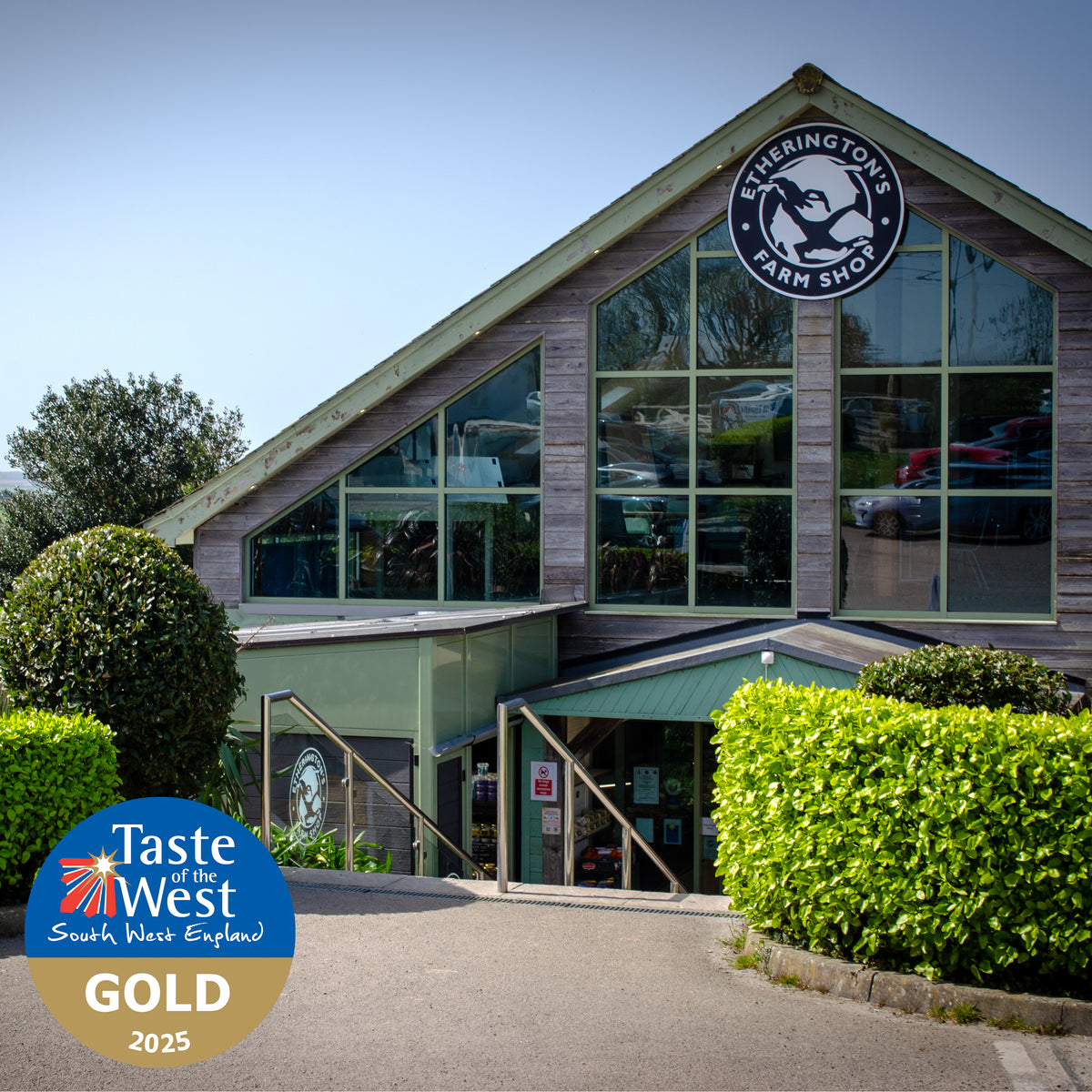 Eisherington's Farm Shop exterior with a large gabled building, glass windows, green roof and awning, trimmed hedges, and a Taste of the West Gold 2025 badge in the lower-left corner.