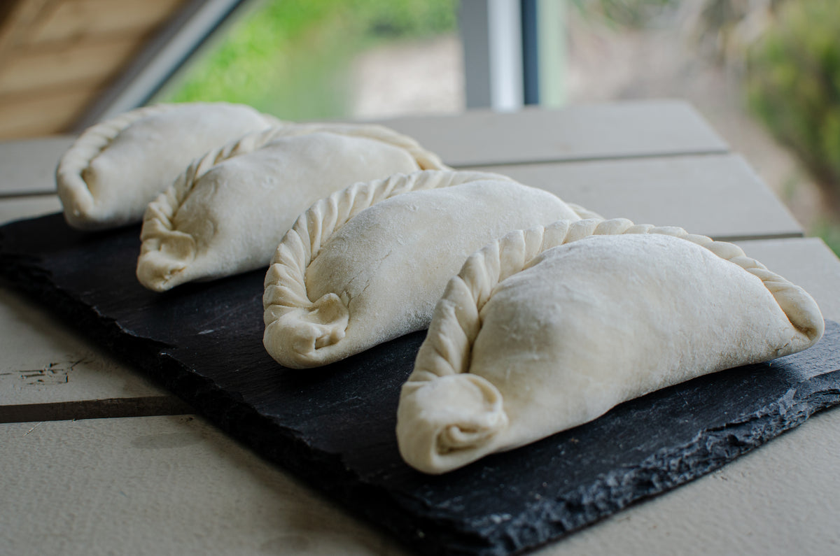 Four uncooked pasties with crimped edges arranged on a black slate serving board on a light wooden table.