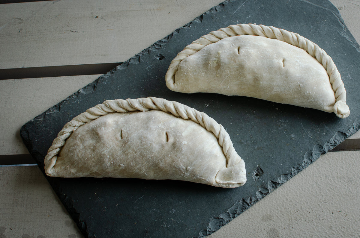 Two uncooked, Cornish pasties resting on a dark slate serving board over a light-colored wooden surface.