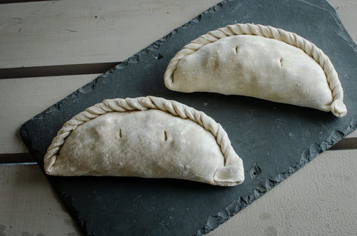 Two uncooked, Cornish pasties resting on a dark slate serving board over a light-colored wooden surface.