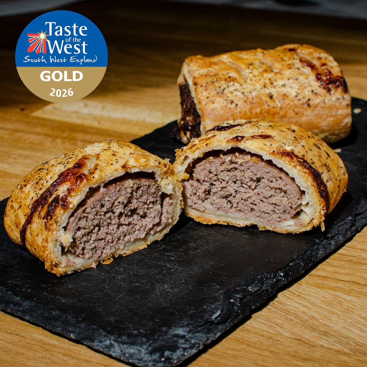 Two lamb and mint sausage rolls on a black slate platter, with one roll cut in half to show the cooked meat filling. A Taste of the West South West England Gold 2026 award badge is in the top left corner.