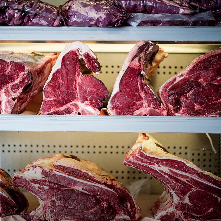 Raw beef steaks and large cuts of red meat displayed on metal shelves in a butchery setting.