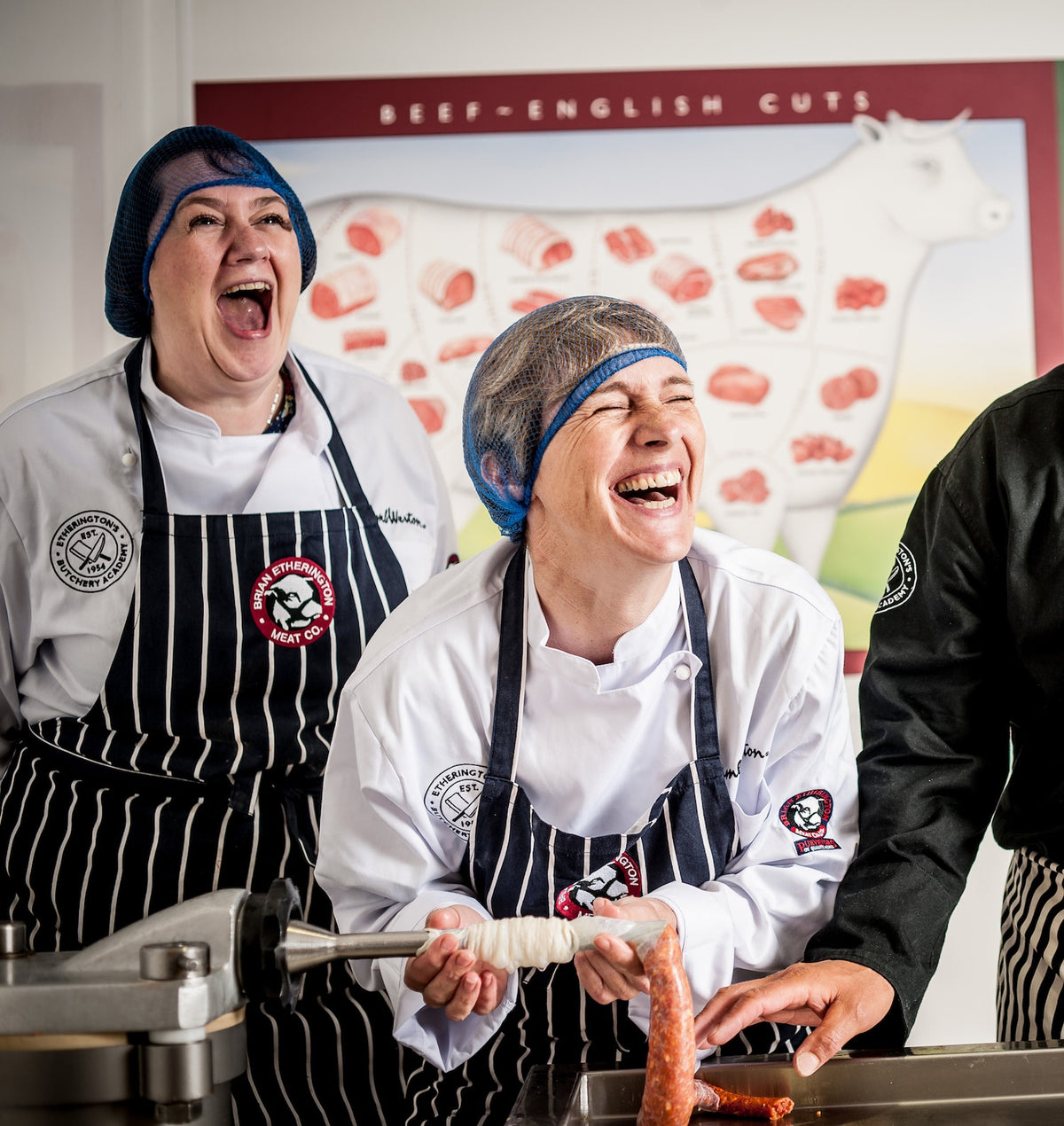 Two people in white chef coats and black-and-white striped aprons laughing while making sausages in a kitchen setting, with a beef cut diagram poster in the background.