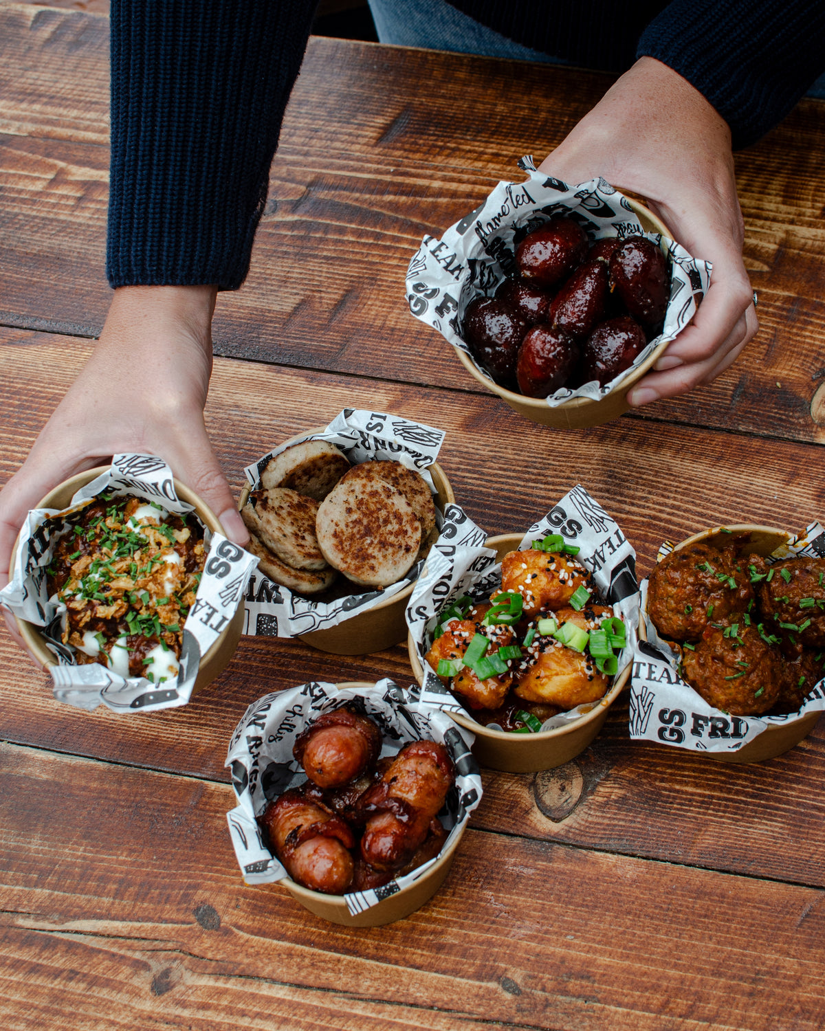 Five small paper-lined bowls of assorted street food arranged on a rustic wooden table. The food includes chorizo bites, BBQ pulled pork, hogs pudding, pigs in blankets, sweet chilli chicken bites and meatballs