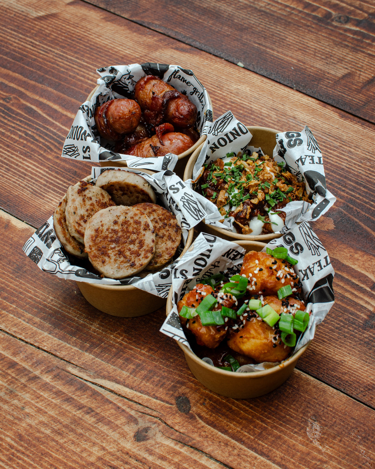 Four small paper bowls of assorted street food arranged on a rustic wooden table. The food includes pigs in blankets, hogs pudding, sweet chilli chicken bites and BBQ pulled pork